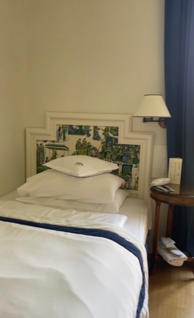 Bright and airy single room at Hotel Elephant in Brixen with crisp white bedding, blue accents, and a decorative illustrated headboard. A bedside table with lamp and telephone sits beside the neatly made bed, offering a comfortable base before hiking Alta Via 2 in the Dolomites.