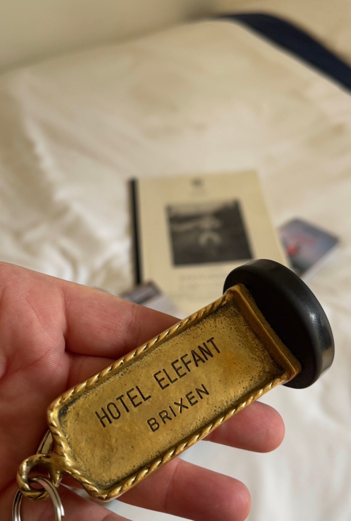 Hand holding a vintage brass hotel key fob engraved with HOTEL ELEFANT BRIXEN in black lettering. A white hotel bed and a book rest softly out of focus in the background, introducing Hotel Elephant in Brixen at the start of an Alta Via 2 trip. Image 2 Hand holding a vintage brass hotel key fob engraved with HOTEL ELEFANT BRIXEN in black lettering. A white hotel bed and a book rest softly out of focus in the background, introducing Hotel Elephant in Brixen at the start of an Alta Via 2 trip.