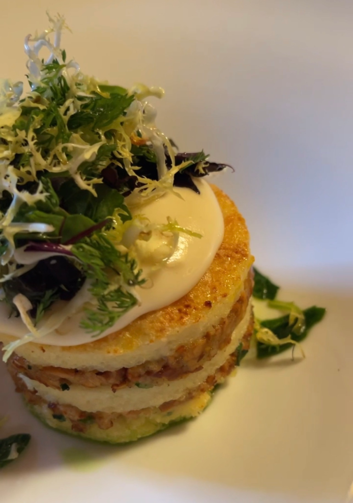 Plated gourmet appetizer at Hotel Elephant in Brixen featuring layered potato rounds with a savory filling, topped with a creamy sauce and fresh mixed greens. The elegant presentation highlights the refined dining experience before setting off on Alta Via 2 in the Dolomites.