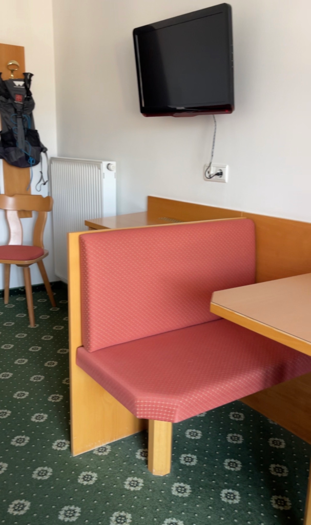 Small seating area at Hotel Trenker featuring a red upholstered bench, wooden table, wall mounted television, and green patterned carpet. The practical layout offers a functional space to organize gear before hiking Alta Via 1.