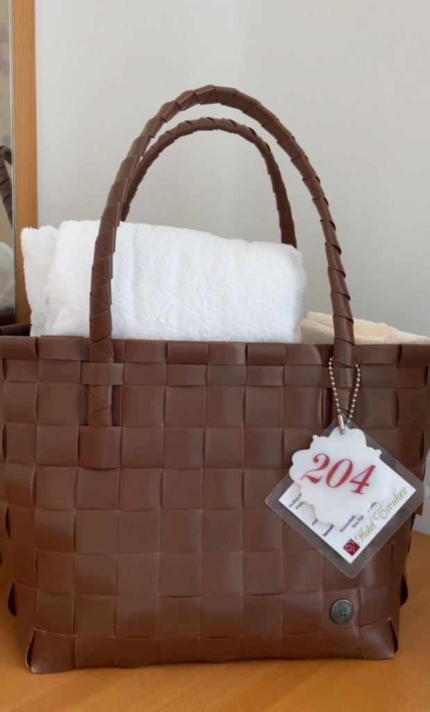 Brown woven spa tote bag with white towels inside and a tag labeled 204 and Hotel Trenker attached to the handle. The provided spa bag highlights the relaxing amenities available before or after hiking Alta Via 1 in the Dolomites.