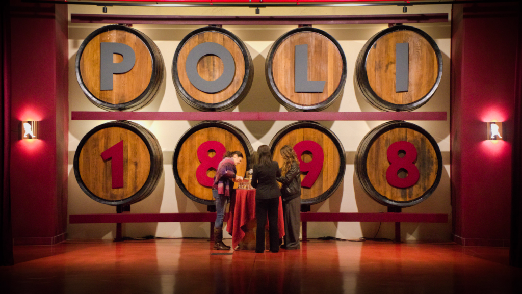Three women sample drinks at a tasting table beneath large wooden barrels that spell out "POLI 1898" at Poli Distilleries in Bassano del Grappa. The warm lighting and vintage decor highlight the historic atmosphere of this grappa-tasting experience, a top day trip from Vicenza.