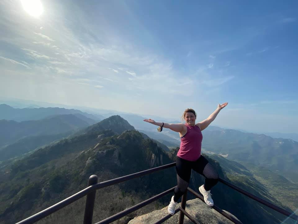 Woman with pink tanktop and black leggings has arms outstretched over mountain top in Songnisan National Park in Korea