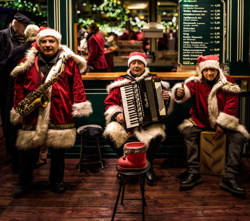 Three men in santa suits sitting down playing instruments while smiling. One is holding a saxophone, middle one is holding an accordion and the last one is drumming on a box. 