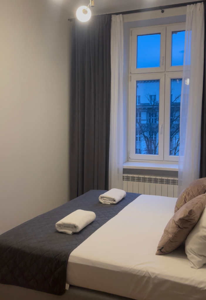 White walled bedroom with a window and navy blue curtains with a bed that has two towels on it and several pillows in navy blue and yellow