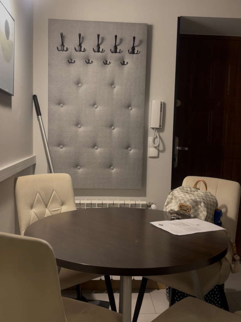 a brown table with a white piece of paper on it and 2 leather bound white chairs with a wall that has cleaning supplies hanging from it