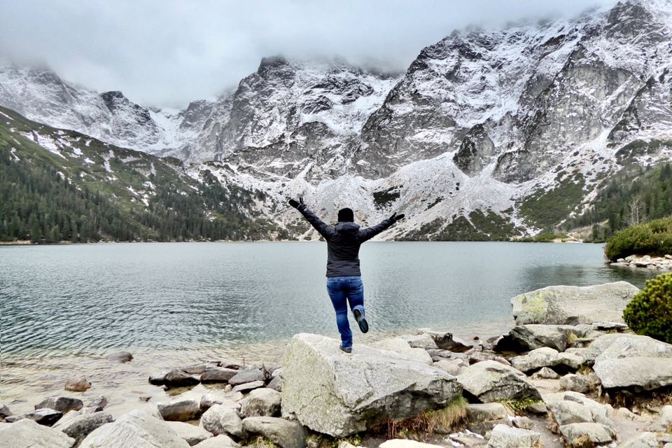 Woman with arms outstretched in a jacket and black hat with blue jeans in front of mountains at Morskie Oko