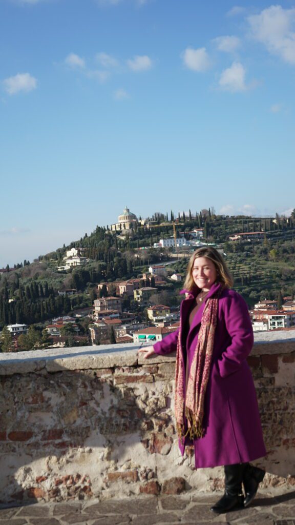 Woman with pink coat and scarf standing in front of a scenic spot in Verona Italy