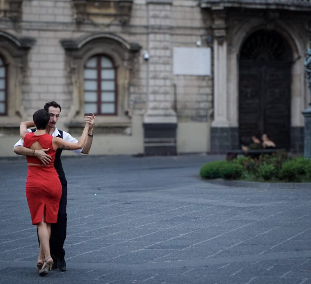 a woman wearing a red dress dances with a man wearing a suit in a piazza in Catania Sicily