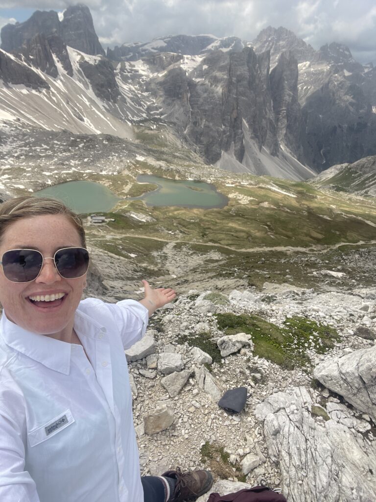 woman smiling at camera with outstretched arms with the mountains behind her