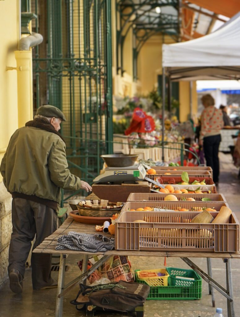 a man in a green jacket and blue jeans stands next to a fruit he is selling at a market