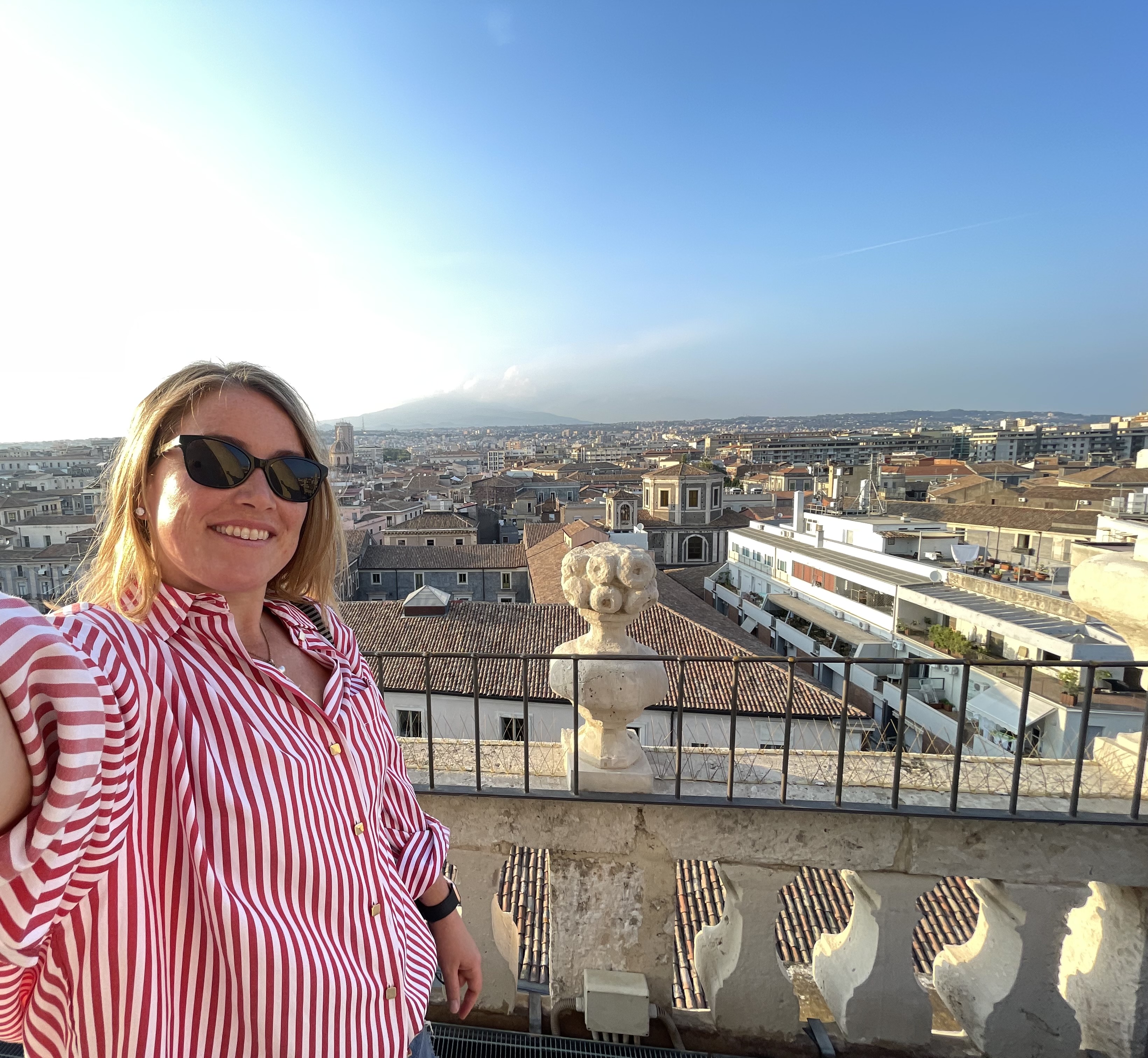 Woman with striped shirt on rooftop with view of Mt. Etna in back against blue skies