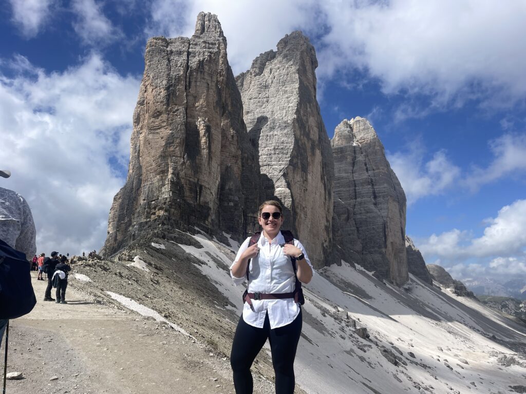 Woman with a white long sleeve shirt, black pants and a backpack standing in front of Tre Cime