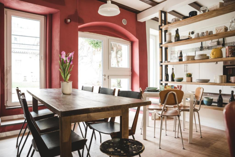 Bright and stylish shared dining area at Hostello in Veronetta, featuring a rustic wooden table with black chairs, open shelving stocked with kitchenware, and vibrant red walls framing large windows.