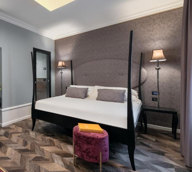 Elegant hotel room in Verona featuring a four-poster bed with neutral bedding, velvet magenta armchairs and ottomans, dark wood herringbone floors, and soft lighting from classic standing lamps.