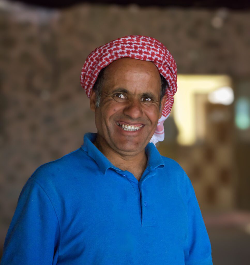 a man smiles at the camera in a blue shirt and red and white keffiyeh