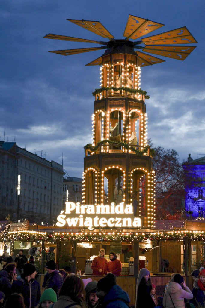 Christmas Pyramid with the words piramida swiateczna on the front against a blue sky with passerbys enjoy the Poznan Christmas Market