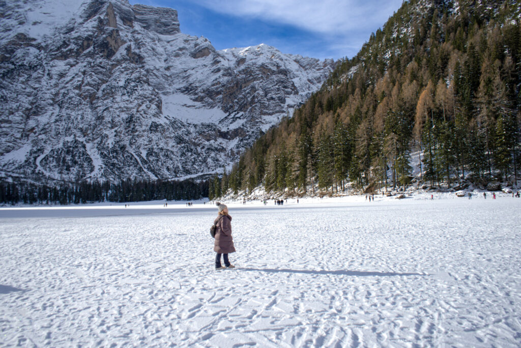 A woman in a long purple coat and gray knit hat walks on the snow-covered shore of Lago di Braies with her brown dog on a leash. This quiet moment captures one of the best things to do in the winter in the Dolomites for nature lovers and pet owners.