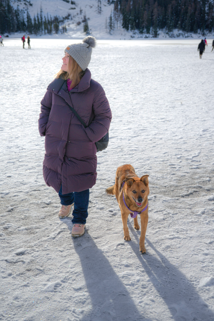 A woman in a long purple coat and gray knit hat walks on the snow-covered shore of Lago di Braies with her brown dog on a leash. This quiet moment captures one of the best things to do in the winter in the Dolomites for nature lovers and pet owners.