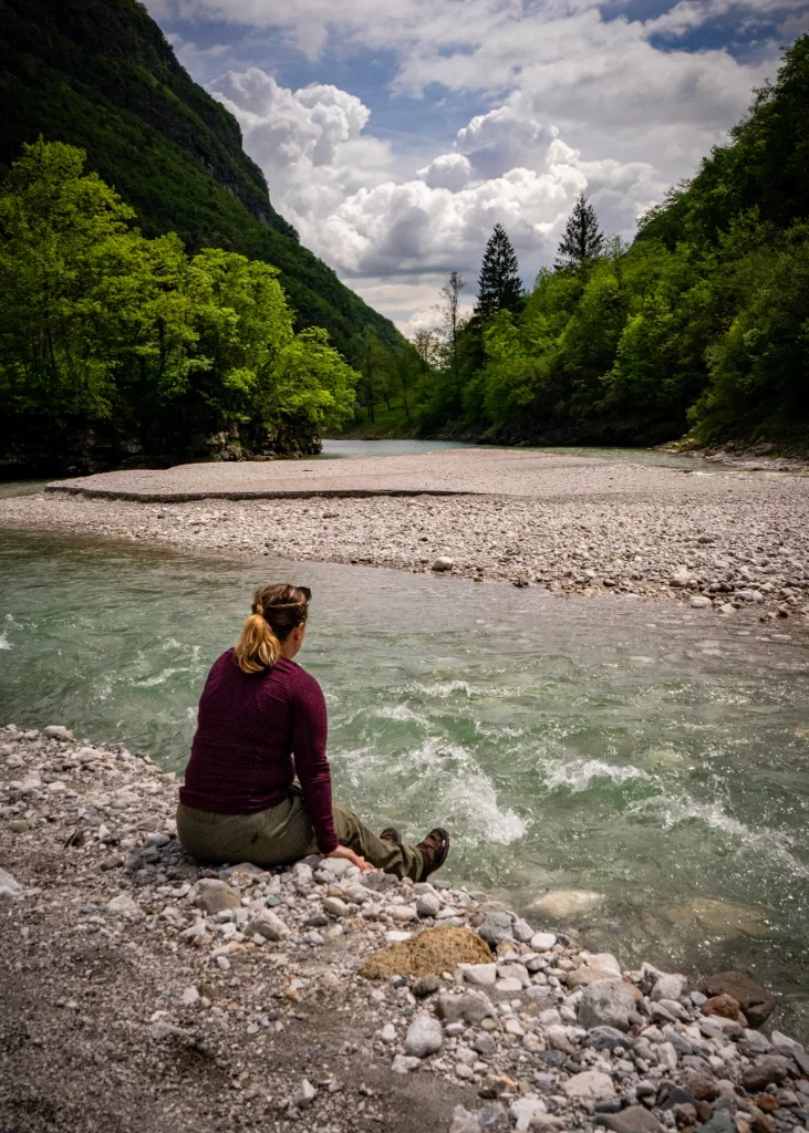Woman sitting on the side of a river  in belluno near the Alta Via 1 with a red long sleeve top and green pants with hiking boots on. 