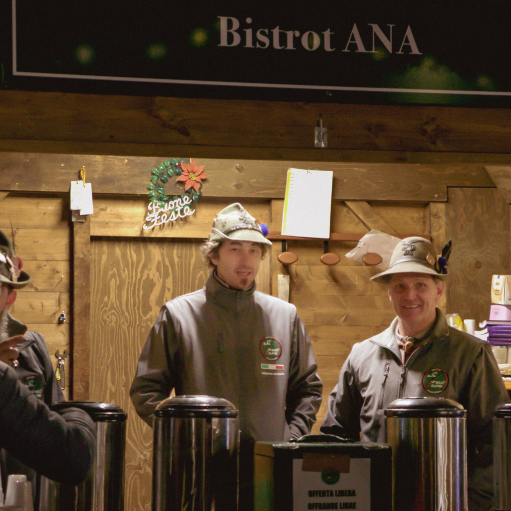two men smile at the camera from the alpini stall in aosta, wearing traditional alpini caps with feathers in their cap handing out vin brule at the Aosta Christmas Market