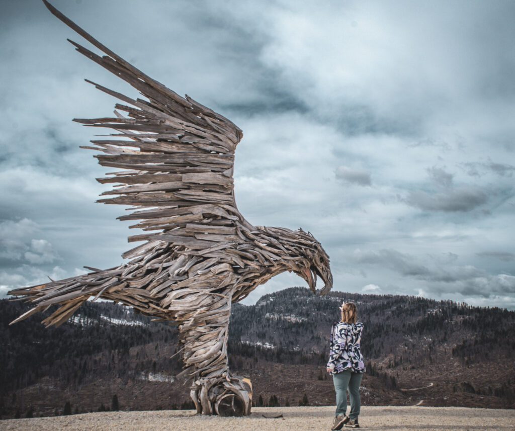 Woman Reaching for Bird made out of wood in an alpine setting against a blue sky
