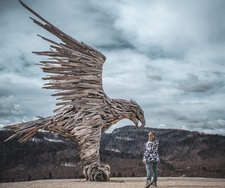 Woman Reaching for Bird made out of wood in an alpine setting against a blue sky
