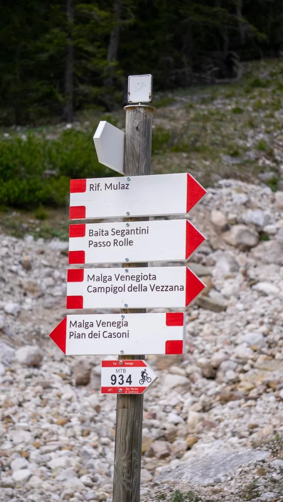 A wooden signpost with red-and-white directional markers points toward Rifugio Mulaz, Baita Segantini, Malga Venegiota, and Malga Venegia in Val Venegia. A key trail junction for day hikers exploring the Dolomites.