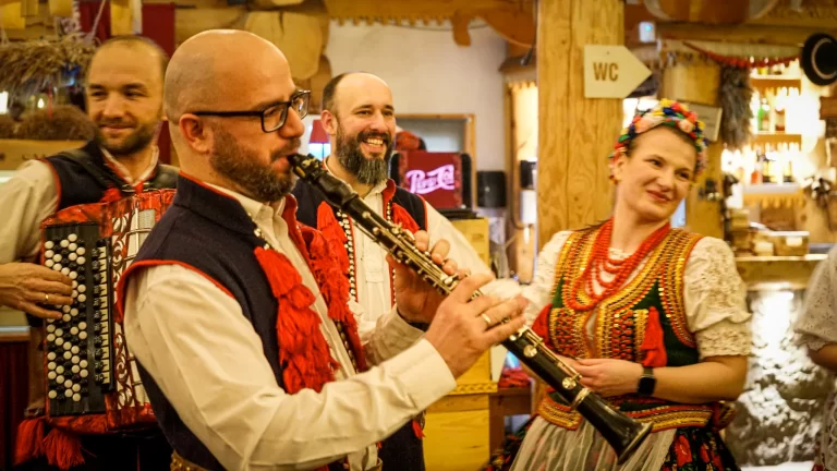 Man blowing into an oboe and wearing traditional polish clothing with red and black designs. Two people in the background look on at Traditional Polish Folk Show Krakow