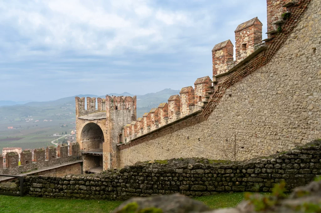 A panoramic view of the crenellated stone wall of a historic castle, curving along the hillside with scenic countryside and rolling hills in the background.