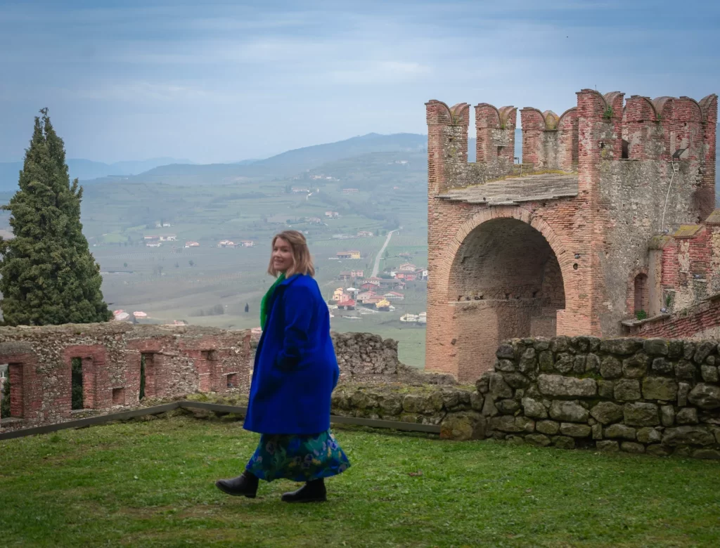 A woman in a bright blue coat and floral skirt walks across a grassy area atop a stone fortress, with a scenic valley and castle tower behind her.