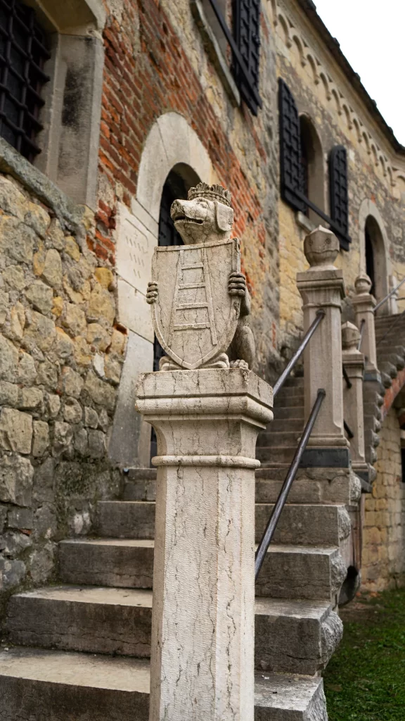 A close-up of a carved stone post topped with a crowned lion holding a shield engraved with a ladder, in front of a historic stone building with black window shutters and a stone staircase at Castello di Soave
