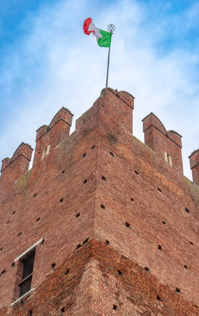The corner of a tall medieval brick tower with crenellated battlements, flying the Italian flag against a partly cloudy sky.