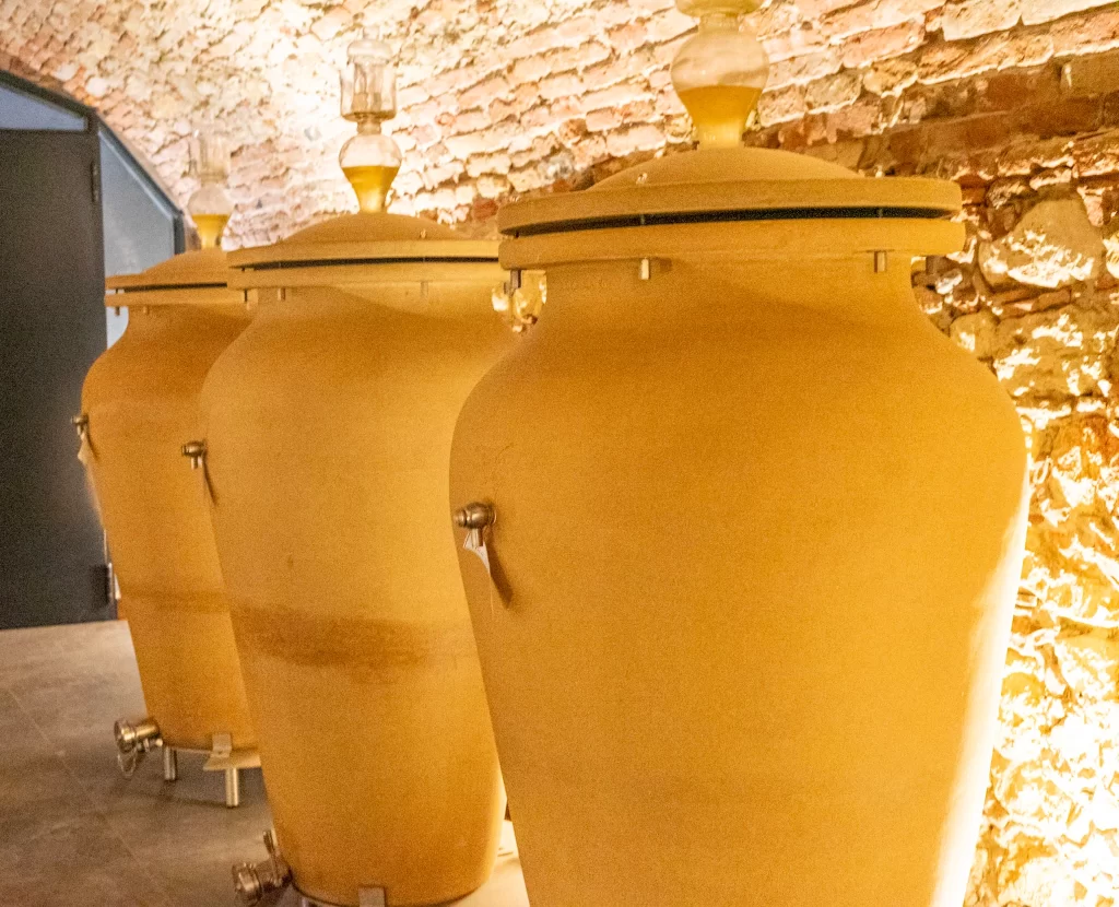 A warm-toned wine cellar with large terracotta amphorae lined up against a brick wall, each fitted with a spigot and glass airlock for fermentation.