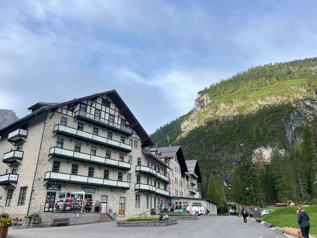 Hotel Lago Di Braies, with its sone facade and beautiful green detail. In the bottom corner a woman walks along the road with a mountain in the background. 