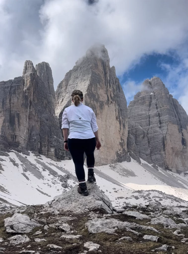Woman with white top and black pants stepping on a rock in front of the Drei Zinnen or Tre Cime di Lavaredo.