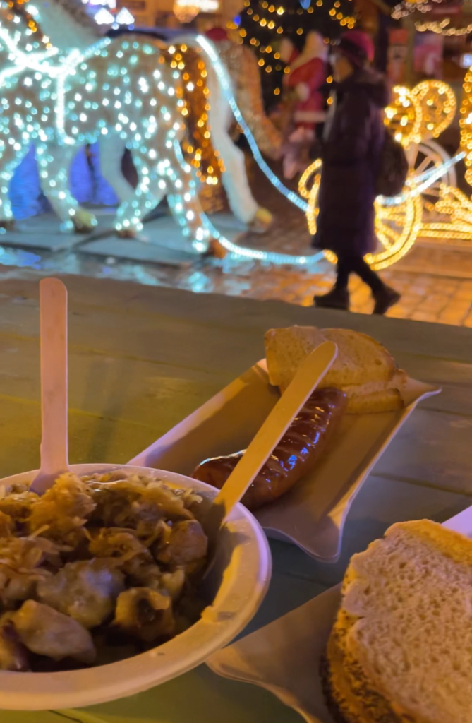 Bigos or hunters stew which is sauerkrat, kielbasa and onion in a bowl with a fork. Kielbasa and bread suround it with christmas market lights in the back