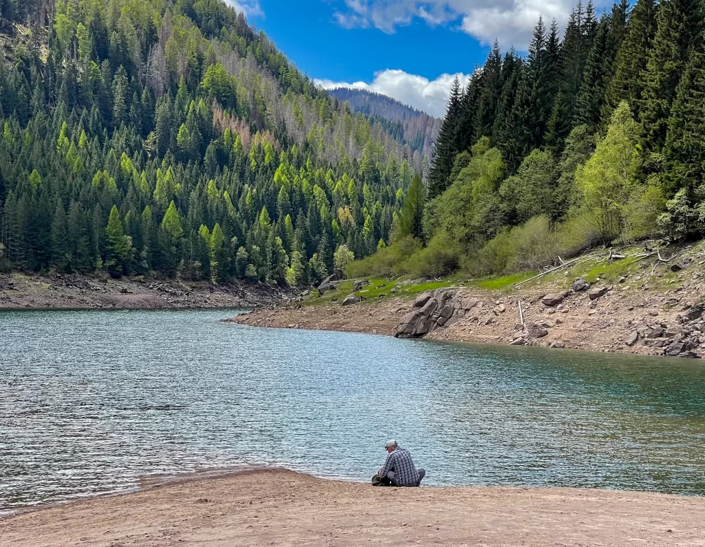 A Man with a hat sitting on the shore of lago di paneveggio set against blue sky and blue water in the Pale Di San Martino in the Italian Dolomites