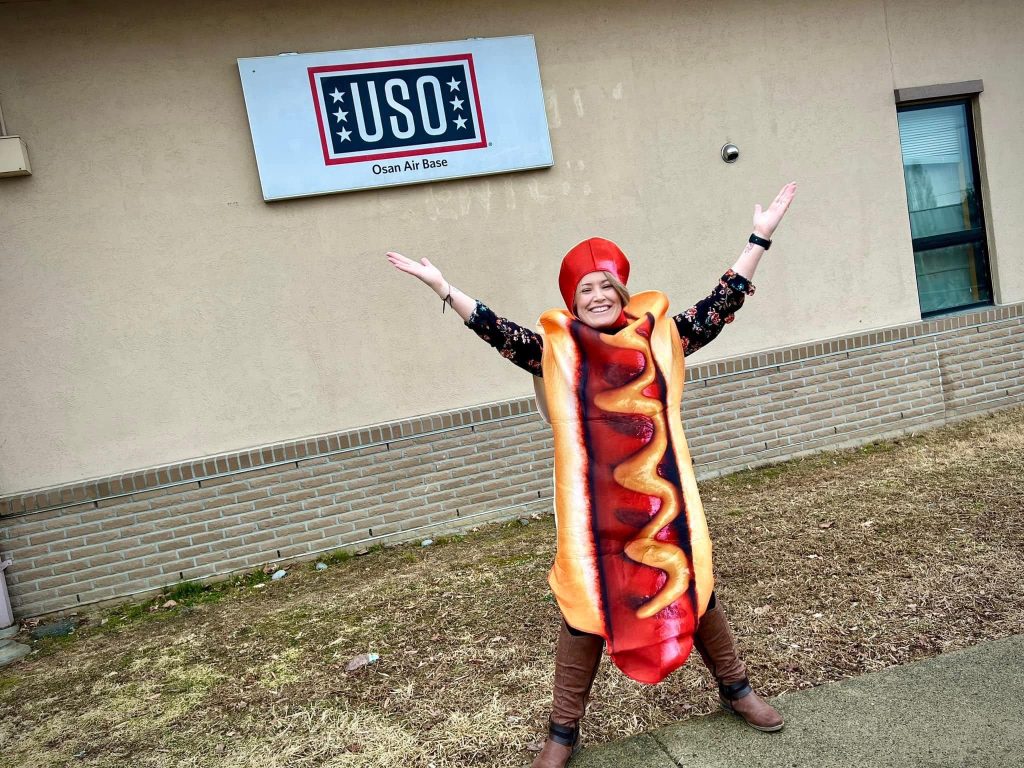 Woman in Hotdog costume in front of a USO sign holding arms up in the air at Osan Air Base in Korea