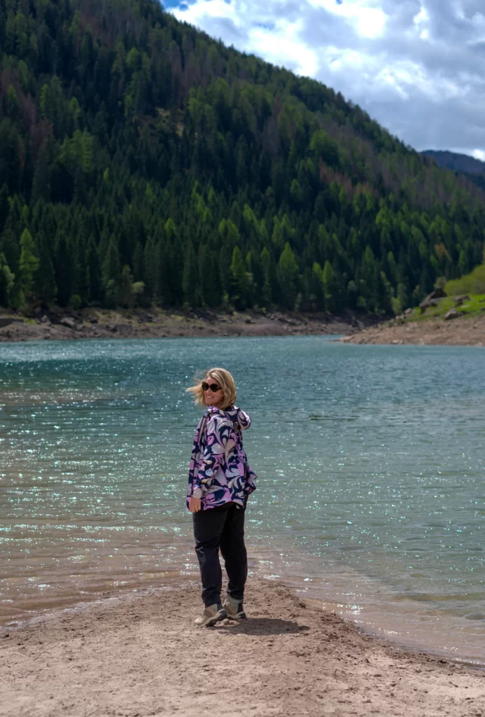 woman standing in front of a lake in the Paneveggio Forest in The Pale Di San Martino in the Dolomites with a blue and purple jacket on and black pants. The sky is blue reflecting on the water of Lago di Paneveggio