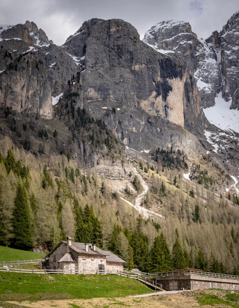 A scenic mountain hut in Val Venegia, surrounded by green meadows and pine forests beneath the towering cliffs of the Pale di San Martino in the Dolomites. An inviting stop along one of the best easy day hikes in the region, with dramatic alpine views and patches of spring snow on the peaks above.