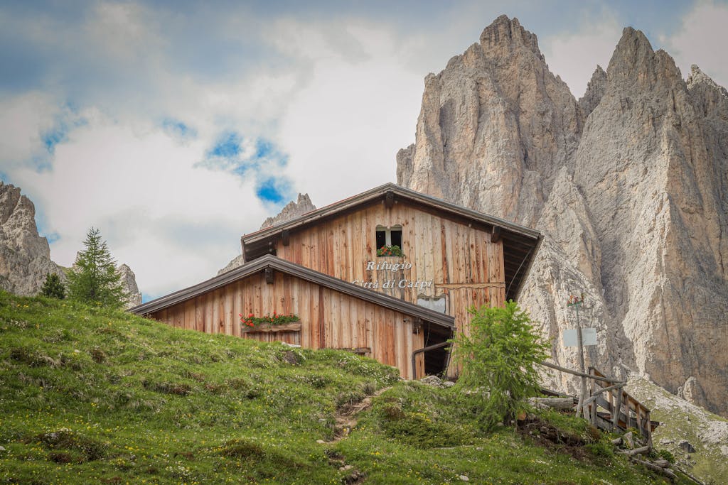 Rustic cabin amidst scenic mountain peaks, lush greenery, and clear skies in the Dolomites.