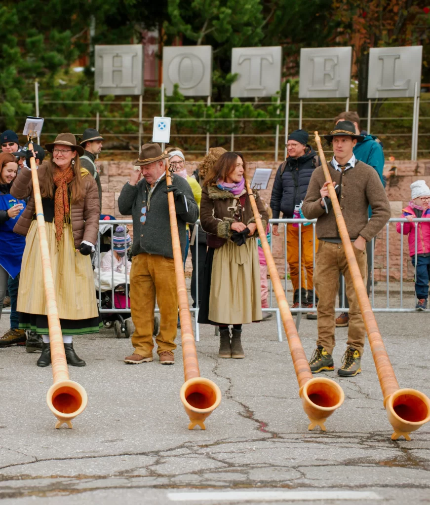 5 people blowing into their traditional alpine horns that are long and brown. They are each wearing traditional alpine clothing