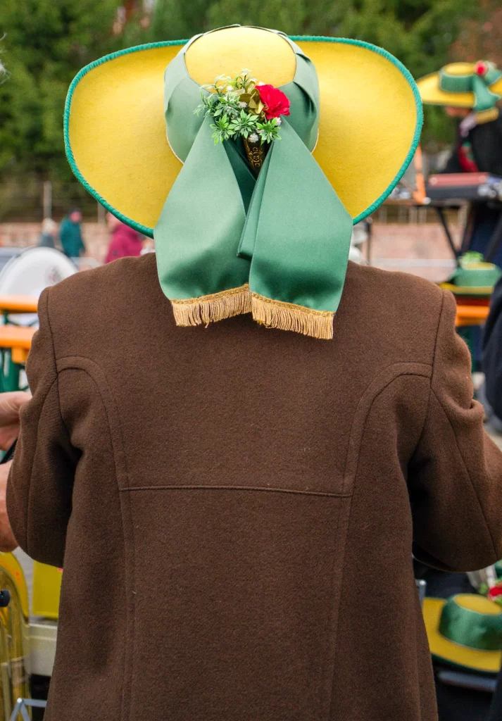 The back of a person who is wearing a yellow hat with flowers and a green ribbon and brown jacket at the Almabtreib in Compatsch, Alpe Di Siusi
