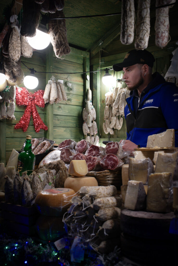 Man in a lit up green stall selling cured meats that are stacked high