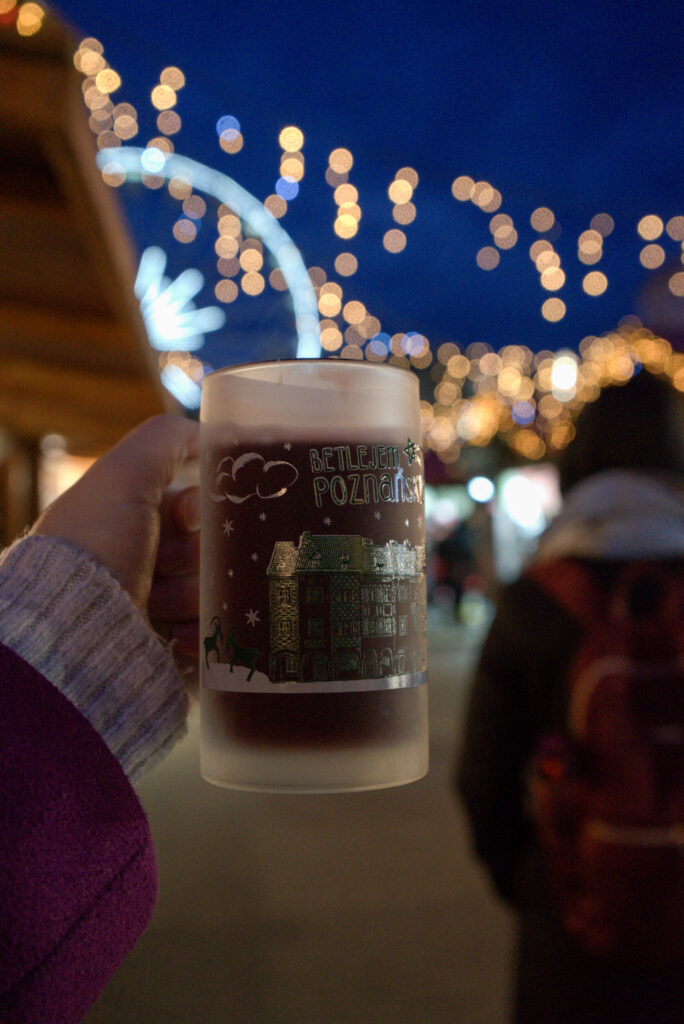 A hand holds up a frosted souvenir mug filled with a warm drink at the Poznań Christmas Market, with “Betlejem Poznańskie” and festive illustrations printed on the cup. In the background, twinkling string lights and a blurred Ferris wheel glow against the evening sky, creating a cozy holiday atmosphere.
