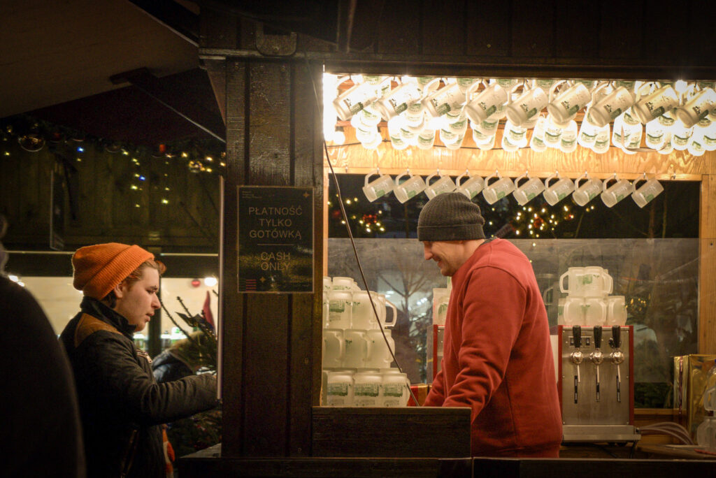 A man in a red hoodie smiles while serving a customer at a cozy Christmas market drink stall. White mugs with green decorations hang above the counter, and a nearby sign reads “PŁATNOŚĆ TYLKO GOTÓWKĄ – CASH ONLY.”