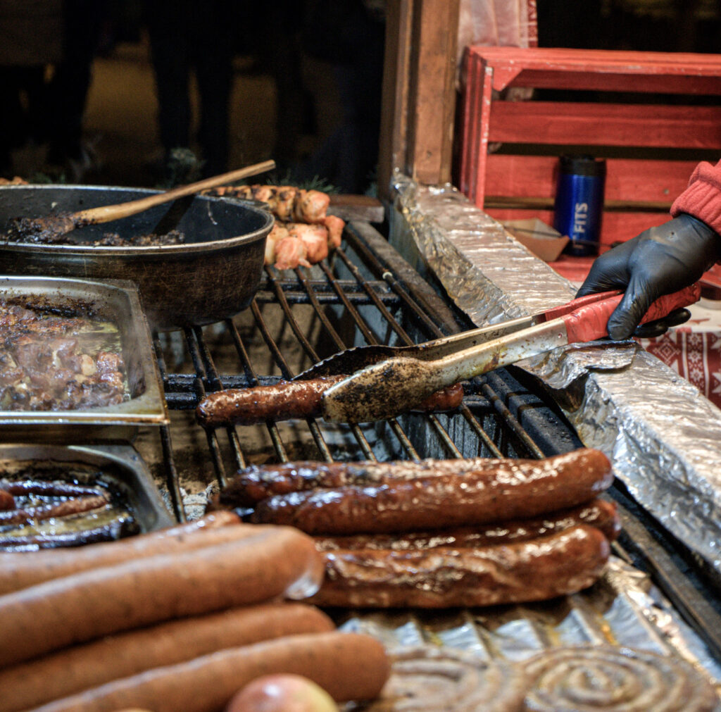 Sausages sizzle on a grill alongside a pot of stewed meat, while a gloved vendor turns one with tongs. The stall setup is rustic, and a bundle of uncooked sausages lies in the foreground.