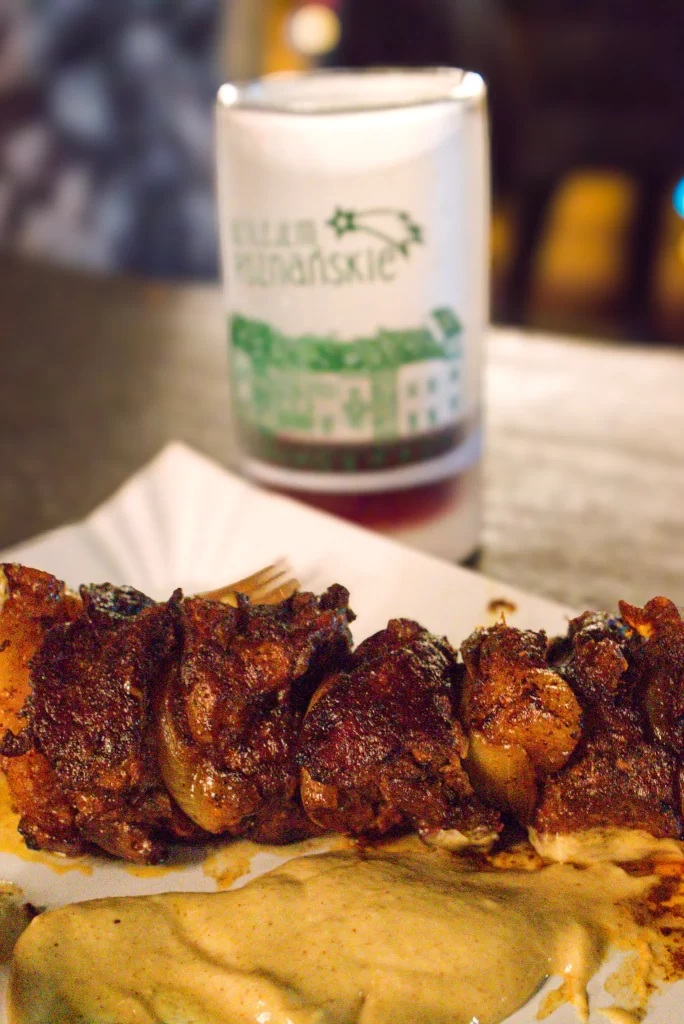 A close-up of grilled meat skewers with a generous coating of seasoning, served on a white paper tray with a dollop of creamy mustard sauce. In the blurred background, a festive white mug with green illustrations and Polish text "KUBEK POZNAŃSKIE" is visible.