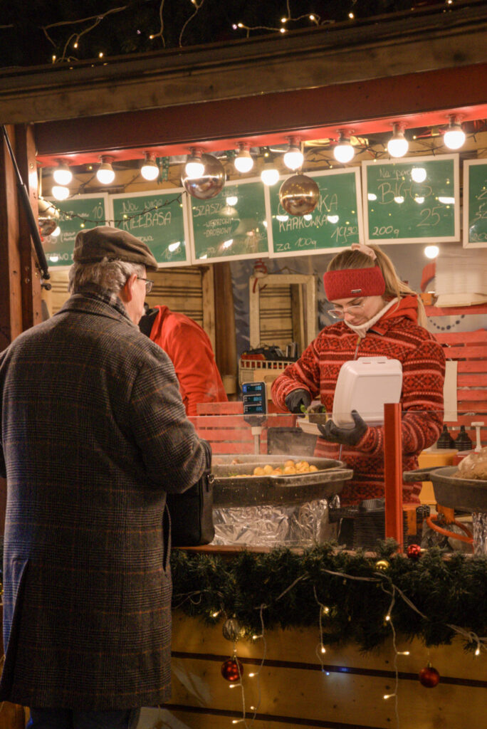A vendor in a red knit sweater and headband serves food into a takeout box at a brightly lit Christmas market stall. A customer in a dark coat and cap waits while handwritten green menu boards overhead list Polish dishes like kiełbasa, szaszłyk, and bigos.